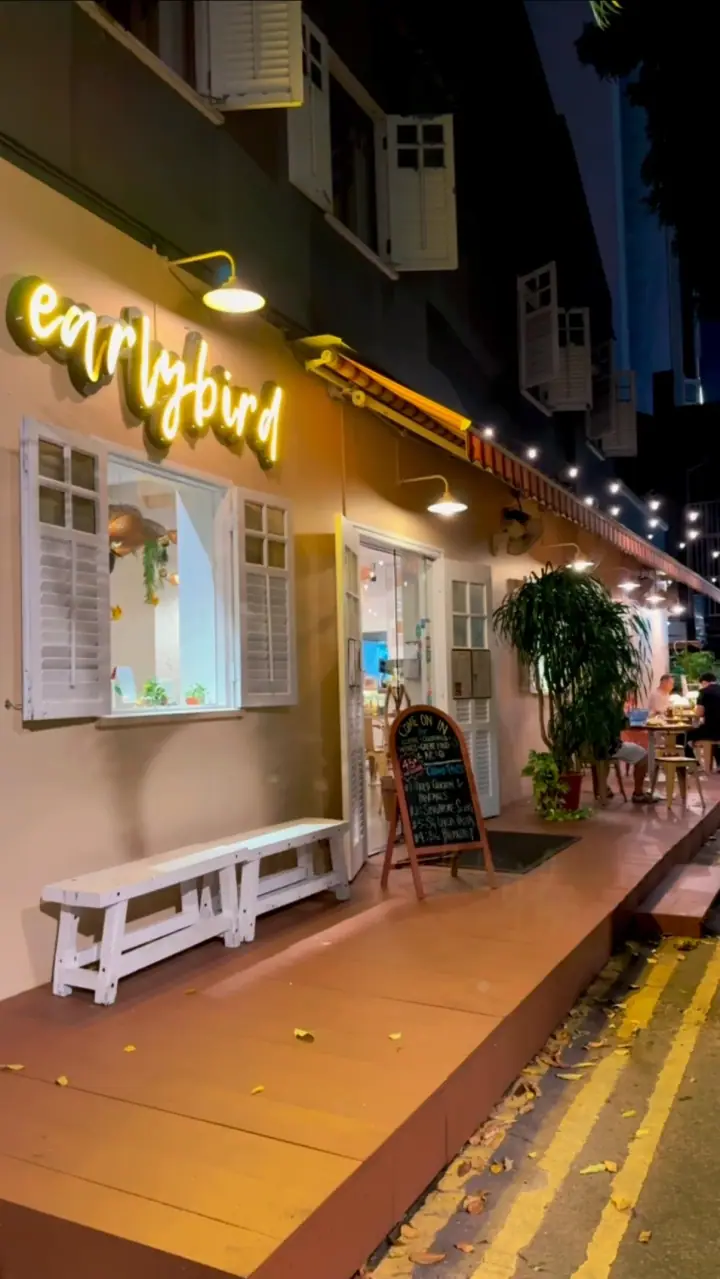 A small dog sitting at the outdoor pavement seating of Earlybird cafe on Jalan Pinang, Kampong Glam, Singapore, with the cafe's open shopfront and a few Kampong Glam shophouses visible behind.