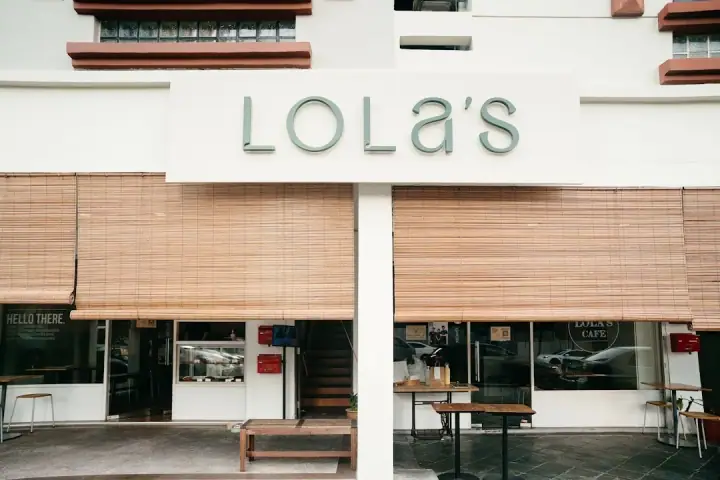 A calm dog resting at its owner's feet inside Lola's Cafe in Kovan / Kensington Park, Singapore, with a ceramic water bowl on the warm wooden floor and soft natural light.