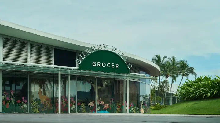 A medium-sized dog sitting beside its owner at Surrey Hills Grocer in VivoCity, Singapore, with the cafe's rainforest theming, hanging plants, animatronic birds, and warm lighting in the background.