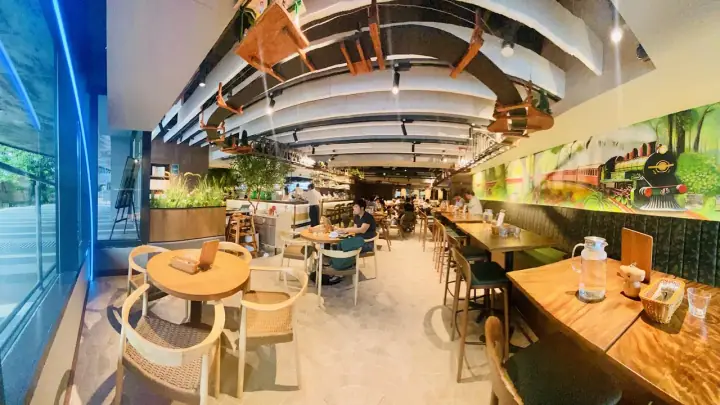 A small dog standing beside its owner at Surrey Hills Grocer in The Woodleigh Mall, Singapore, with the cafe's vintage train-station theming, station-bench seating, and the pet retail corner in the background.