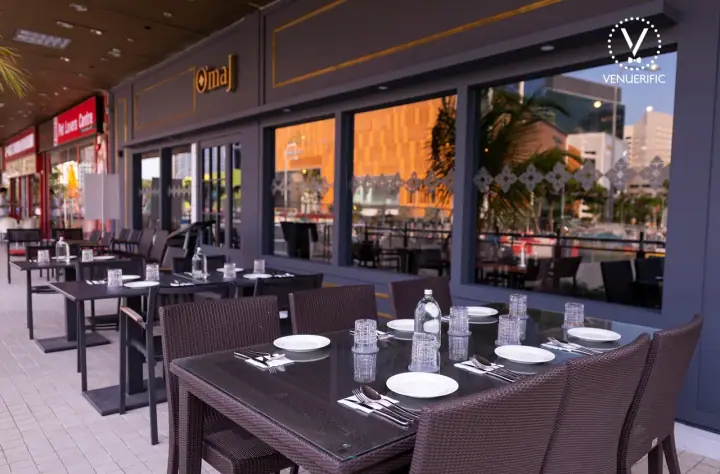 A large dog resting beside its owner at the covered outdoor terrace of Tomahawk Steakhouse at Novena Gardens on Thomson Road, Singapore, with the restaurant's warm interior and steaks on display visible inside.