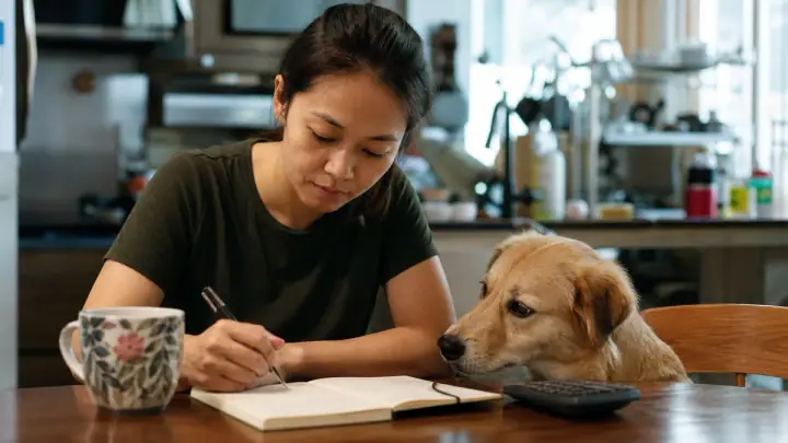 An owner at a kitchen table with a calm dog and an open notebook, working out a household budget